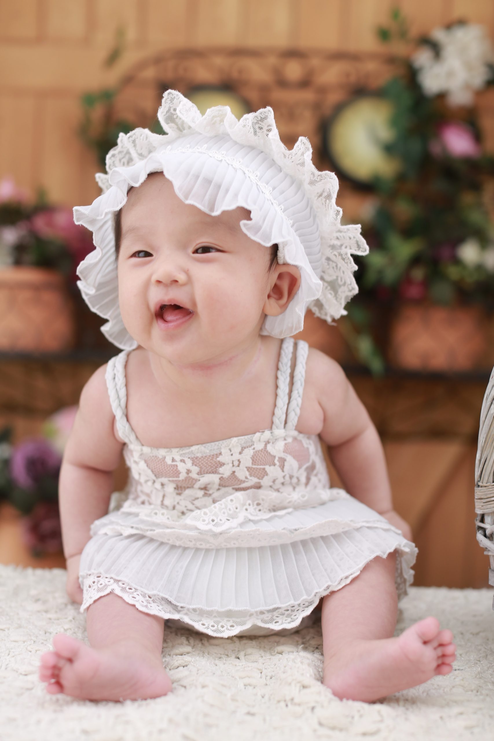 A sitting baby is dressed in a pinkish dress and a frilly lace bonnet.