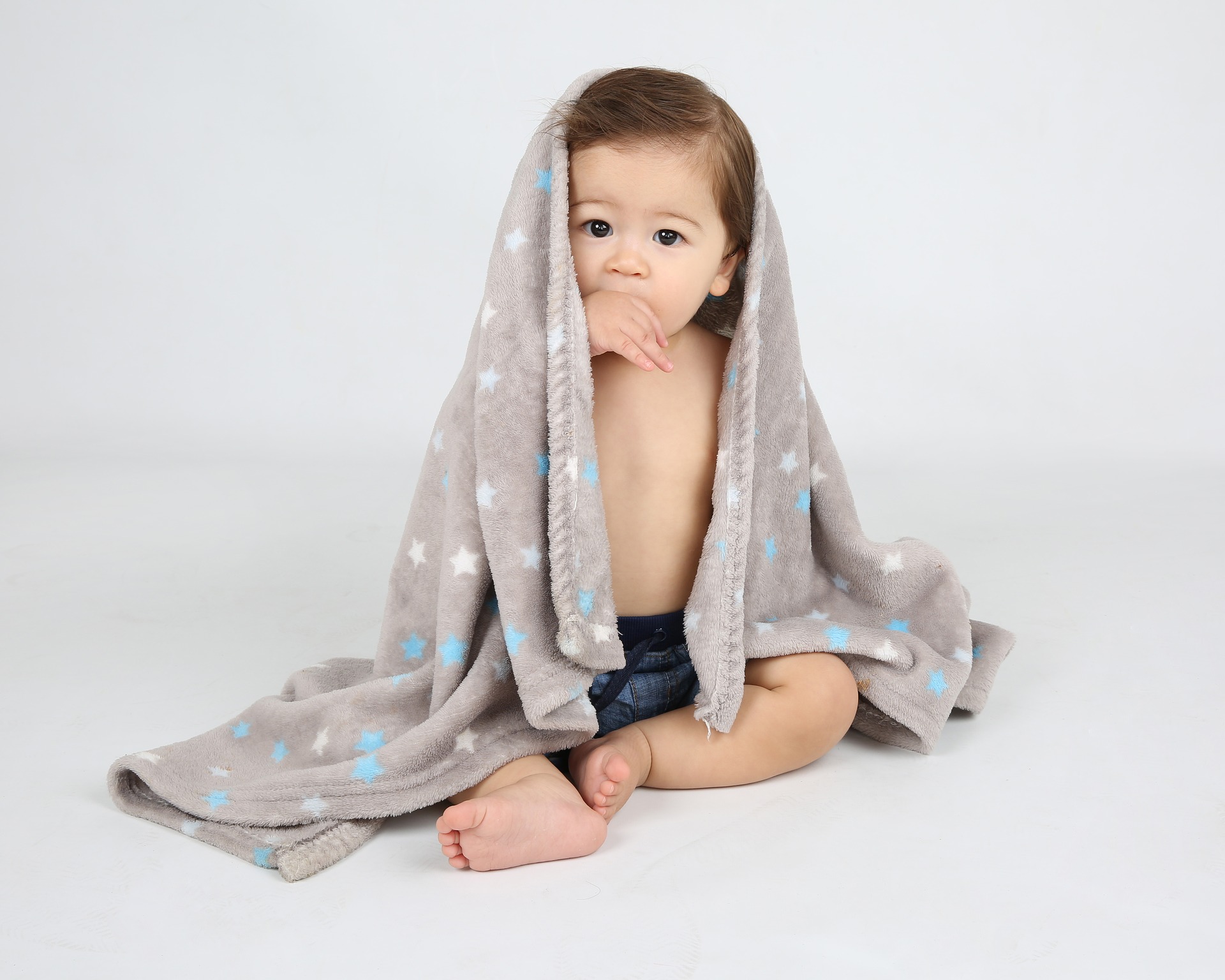A baby sits against a white background while being draped in a grey blanket with different colored polka-dots.