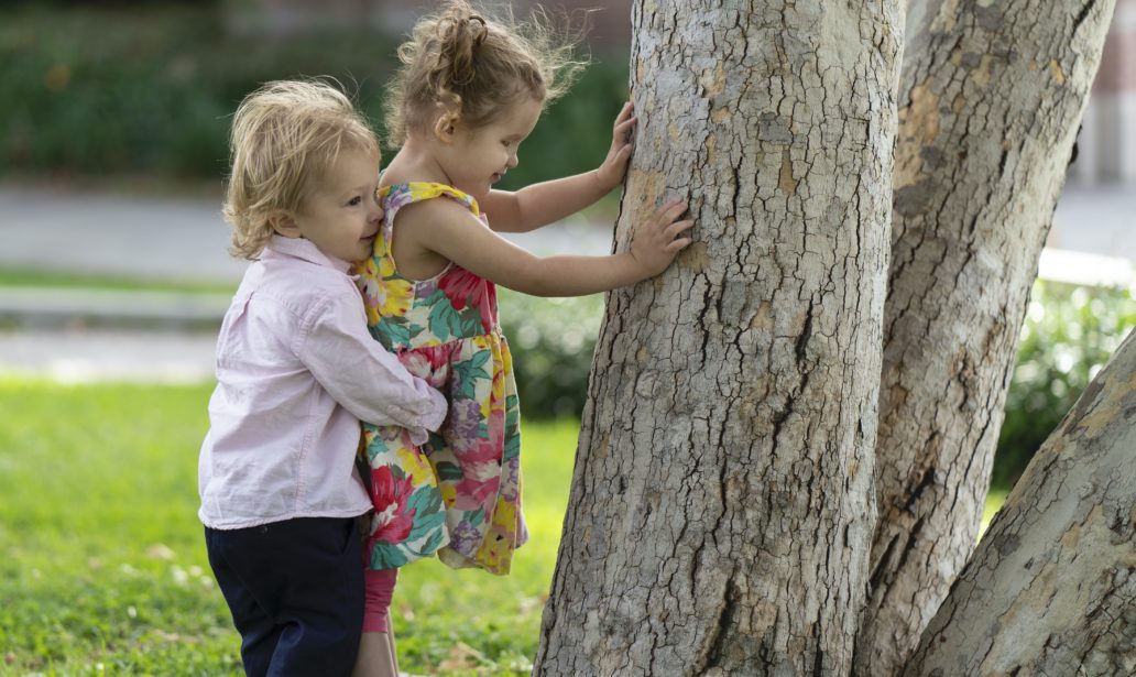 Here, Ellie (wearing a floral print dress and pigtails) explores a tree on the UCLA campus with her hands while her baby brother Sebastian gives her a big hug from behind (he is also blonde and wearing a pink shirt).