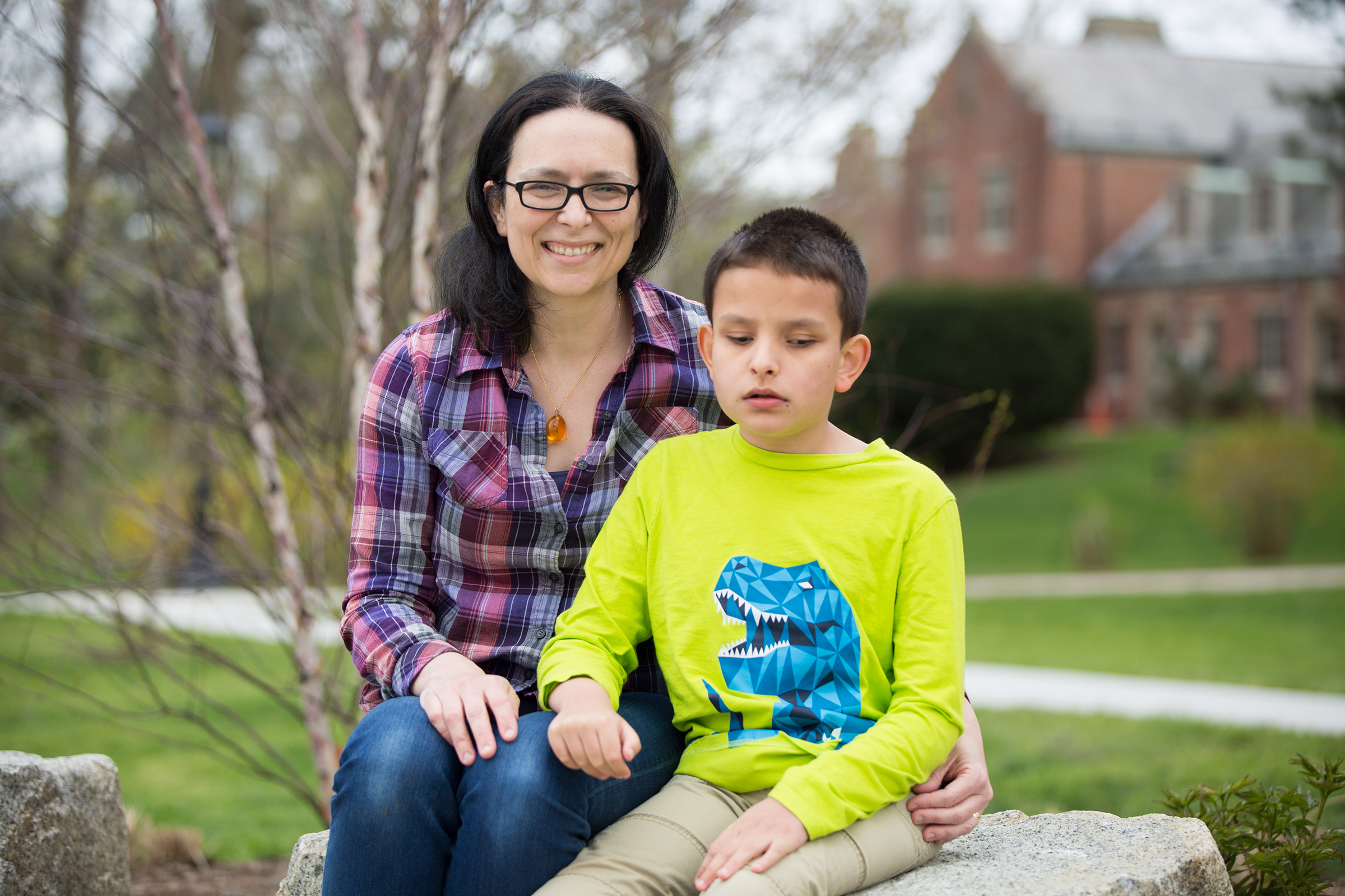 Amber and Ivan are seated on a large rock facing the camera. Amber is smiling, wearing a plaid shirt, jeans, glasses and an amber necklace. She has her arm around Ivan, who wears a green long-sleeve shirt with a blue t-rex and khakis. There are a few trees and buildings in the background.