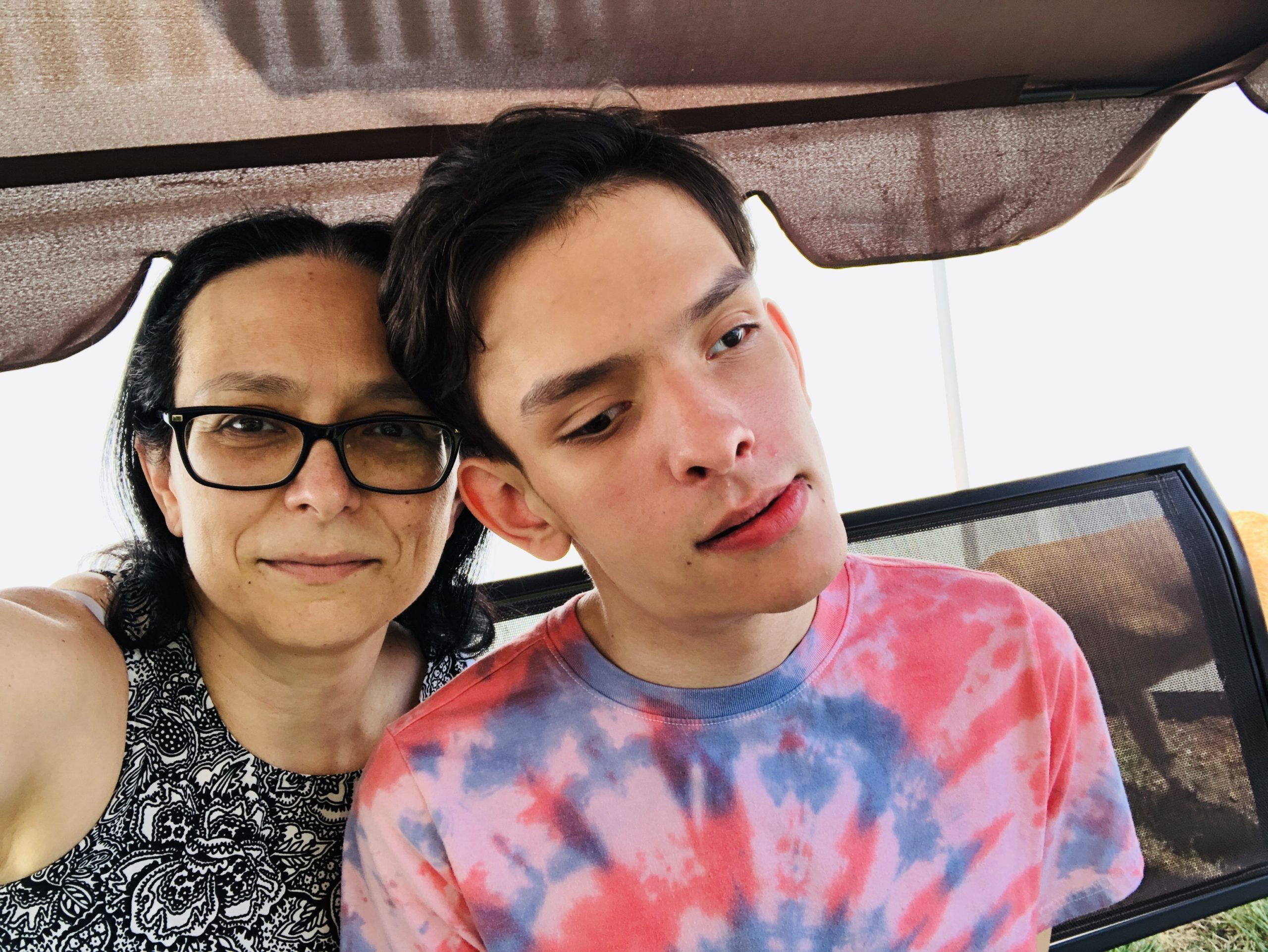 A selfie showing Amber on the left and Ivan on the right. Amber has long, dark brown hair, and wears a black and white patterend shirt and glasses. Ivan has short, dark brown hair and wears a red and blue tie-dye t-shirt.