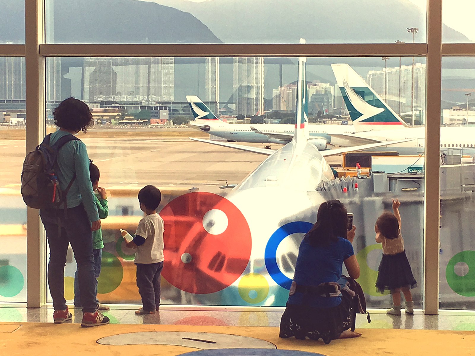 Two mothers and their children stand with their backs to the camera looking out the window at an airport. The family on the left has two children and the family on the right has one toddler, who is reaching up towards the window while the mother takes a picture.