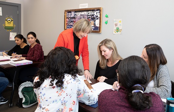 Four women sit around a classroom table while their older female teacher gives them hands on instruction. Two other women look on from a nearby table.