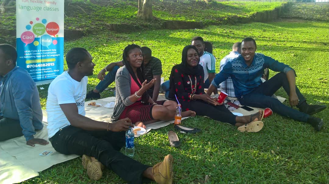 Eight adults sit on a blanket outdoors at the Smarter Language Academy German hangout. They apper to be engaged in conversation.