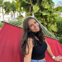 Sophie beams at the camera while sitting on a hammock.. She has tan skin and long brown hair. She wears a black top and jean shorts. There is a leafy tree in the bckground.