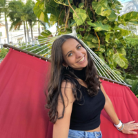 Sophie beams at the camera while sitting on a hammock.. She has tan skin and long brown hair. She wears a black top and jean shorts. There is a leafy tree in the bckground.