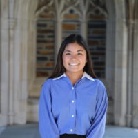 Natalie stands grinning in front of the ornate doors of the Duke chapel. She wears a blue button-down shirt and has dark hair nd brown eyes.