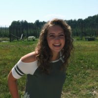 Taylor is laughing and looking at the camera. She wears a grey and white t-shirt, has light skin, and curly light brown hair. The clue blue sky and some grass are visible in the background.