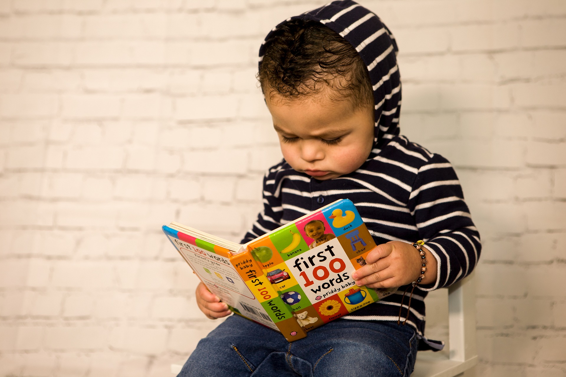 A baby with brown skin and curly black hair sits reading a book with the title, "First 100 words." The baby wears a black and white horizontally-striped hoodie and jeans and has furrowed their brow in concentration.