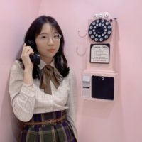 Cai has black hair and glasses. She wears a collared white shirt with a bow at the collar and a brown paid skirt. She stands against a bright pink wall, holding a rotary phone to her ear.