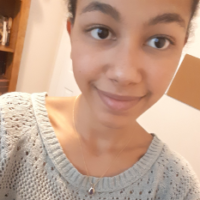 Jasmine smiles at the camera in a close-up selfie. She has natural hair pulled back in a bun, brown eyes, and wears a grey sweater and a necklace.