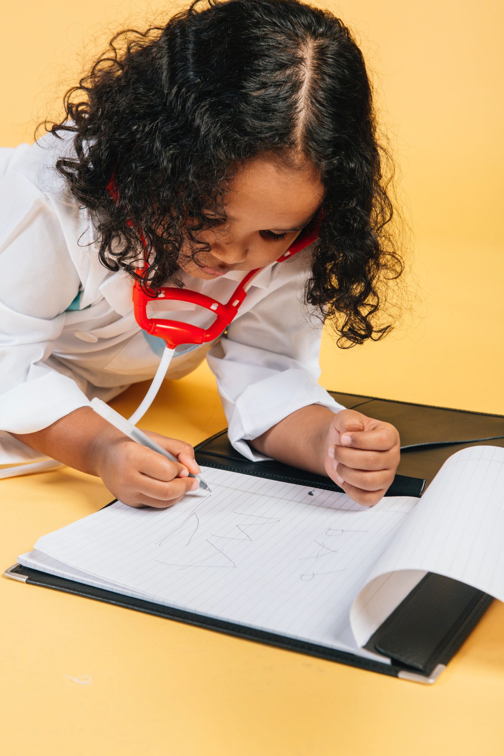 A child with brown curly hair wears a lab coat and a stethoscope and leans over a notebook.