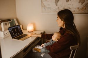 A woman with long dark hair and a maroon turtleneck holds a newborn in a blue onesie on ehr lap as they both sit in front of a laptop, on a video call.