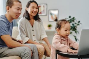 A man and a woman sit behind their toddler, who wears a ruffly pink dress and a little ponytail and is typing on a laptop.