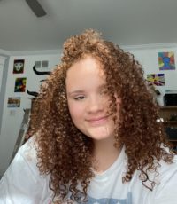 A sunny selfie of Sam smiling. She wears a white tshirt and has curly red hair.