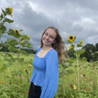 Casey stands in a field of wildflowers with storm clouds in the back.She has light brown hair and a blue shirt.