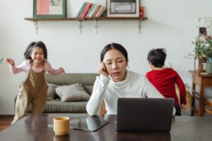 A dark haired woman in a white turtleneck sits in front of a laptop, looking disgruntled, while two children appear to run around yelling in the background.
