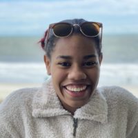 Daisja grins with a beach background, wearing a fluffy jacket and cool sunglasses.