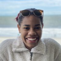 Daisja grins with a beach background, wearing a fluffy jacket and cool sunglasses.