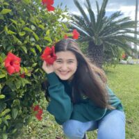 Sarah, a white woman with dark hair, kneels and grins by a hibiscus bush with one of the bright red flowers "tucked" behind her ear.