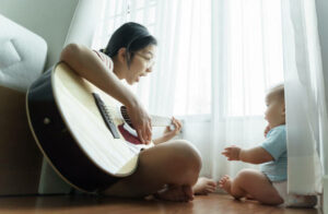 Happy smiling Asian mother and little Cute son sitting on wood floor singing and playing acoustic guitar together.