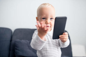 Cute toddler in a stripy shirt holding smart phone. He is sitting on a sofa and is focused on a mobile phone, making an exasperated gesture.