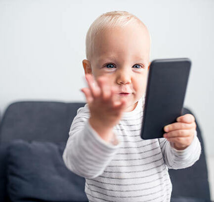 Cute toddler in a stripy shirt holding smart phone. He is sitting on a sofa and is focused on a mobile phone, making an exasperated gesture.