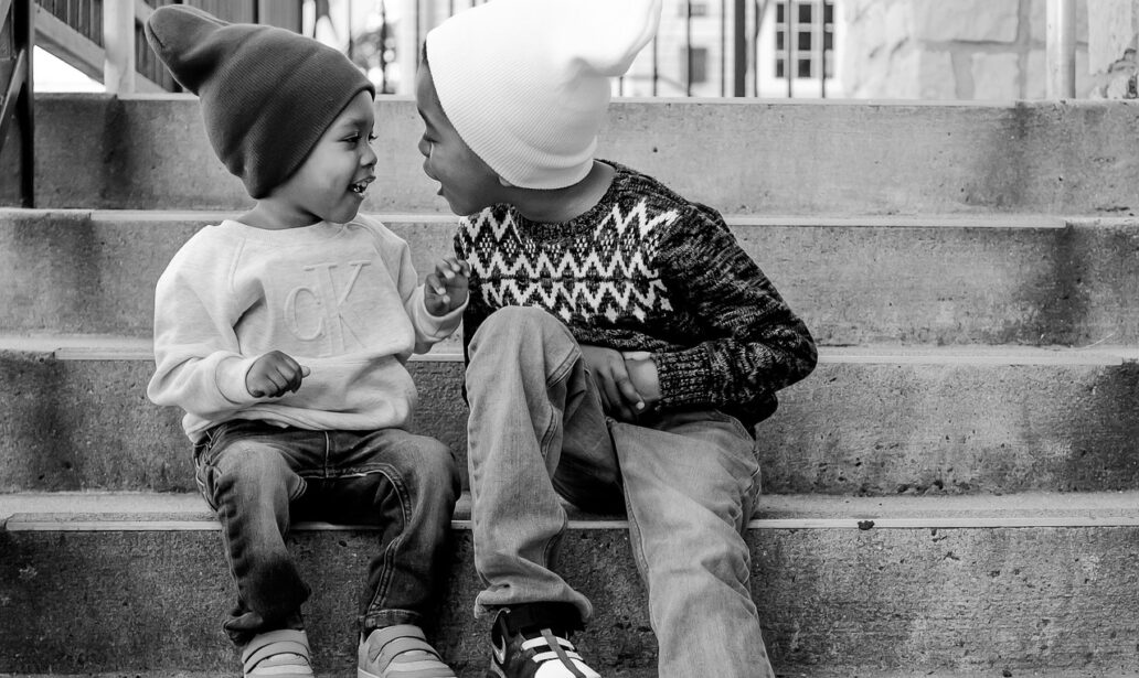 Two young brothers wearing cozy beanies and sweaters sit on the steps grinning at each other, in a high quality black and white image.