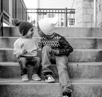 Two young brothers wearing cozy beanies and sweaters sit on the steps grinning at each other, in a high quality black and white image.