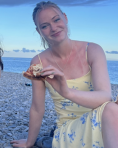 Olivia, a blonde woman wearing a yellow sundress, sits on a beach at dusk and holds a slice of pizza. 